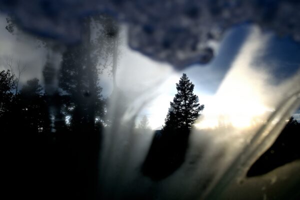 Sunset silhouettes of conifers and aspens peek through frost and ice on a windshield.