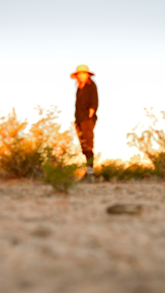 A person stands out of focus with their hands in their pockets, looking at the camera on the desert floor of West Texas, just beyond the morning shadow. This photo was taken in 2021 by American photographer Carson Goldsmith.