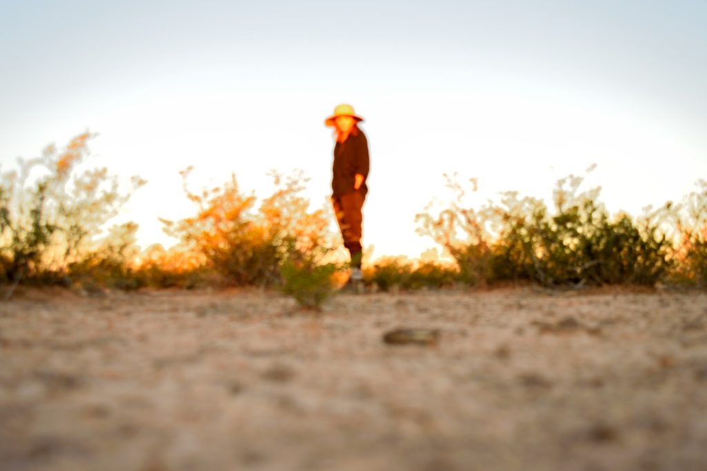 A person stands out of focus with their hands in their pockets, looking at the camera on the desert floor of West Texas, just beyond the morning shadow. This photo was taken in 2021 by American photographer Carson Goldsmith.