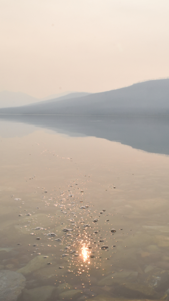 The clear water of Lake McDonald in Glacier National Park reveals various stones and reflects mountains in the distance. The orange Sun shows itself in the midst of water bubbles.