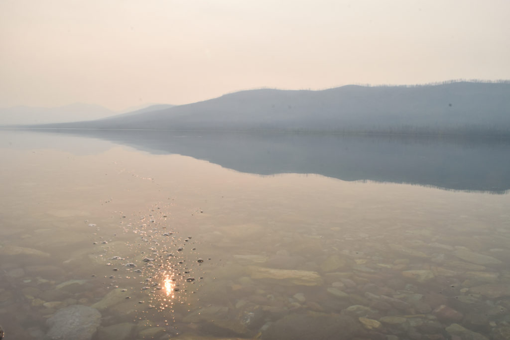 The clear water of Lake McDonald in Glacier National Park reveals various stones and reflects mountains in the distance. The orange Sun shows itself in the midst of water bubbles. 