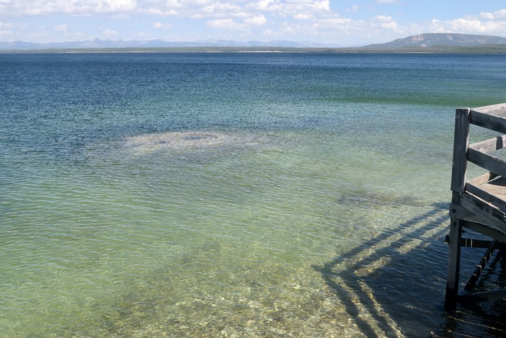 An underwater geyser spout can be seen through the clear green and blue thermal water of Lake Yellowstone. The shadow of a wooden walkway points into the water. Photo by American photographer Carson Goldsmith, captured in 2022.