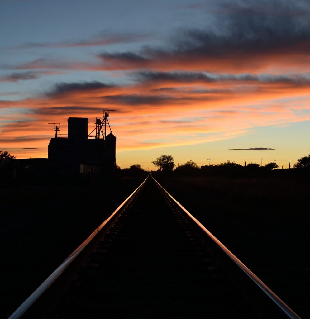 The sillhouette of a building looms over a set of train rails that reflect a vibrant West Texas sunset under peach and slate clouds.