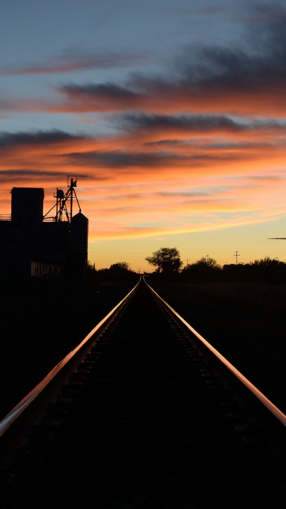 The sillhouette of a building looms over a set of train rails that reflect a vibrant West Texas sunset under peach and slate clouds.