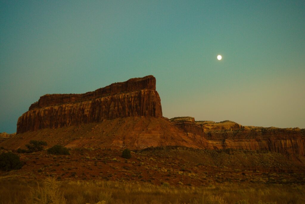 A deep red butte points to the gibbous moon in the dim sky of a Utah evening. 